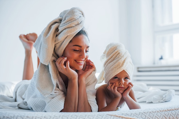 Divirtiéndose. Joven madre con su hija tiene día de la belleza en el interior de la habitación blanca.