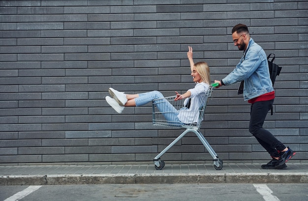 Divirtiéndose y montando carrito de compras Joven elegante con mujer en ropa casual al aire libre juntos Concepción de amistad o relaciones