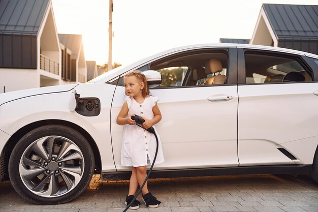 Divirtiéndose Niña de pie cerca de un coche eléctrico con cable de carga en las manos