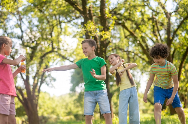 Divirtiéndose. Niños y niñas en edad escolar feliz alegre en ropa brillante jugando con pompas de jabón en el césped en el parque verde