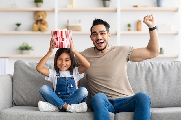 Divirtiéndose. Retrato de feliz padre e hija sentados en el sofá y vitoreando, agitando los puños cerrados, niña sosteniendo una canasta de palomitas de maíz en la cabeza, celebrando el éxito, pasando tiempo libre juntos