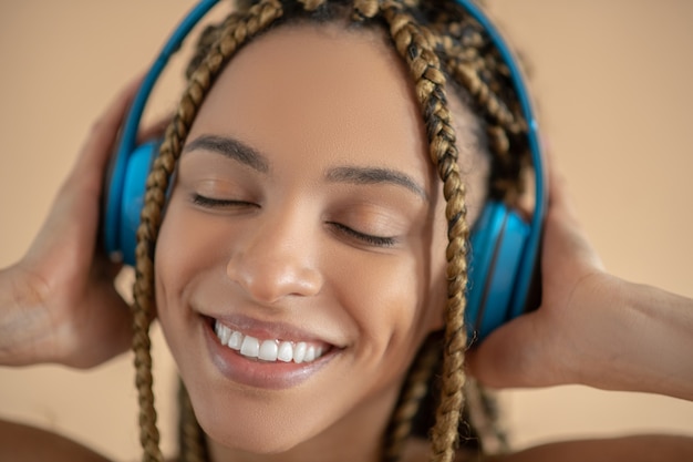 Divirtiéndose. Sonriente joven mujer afroamericana en auriculares azules, escuchando música con los ojos cerrados