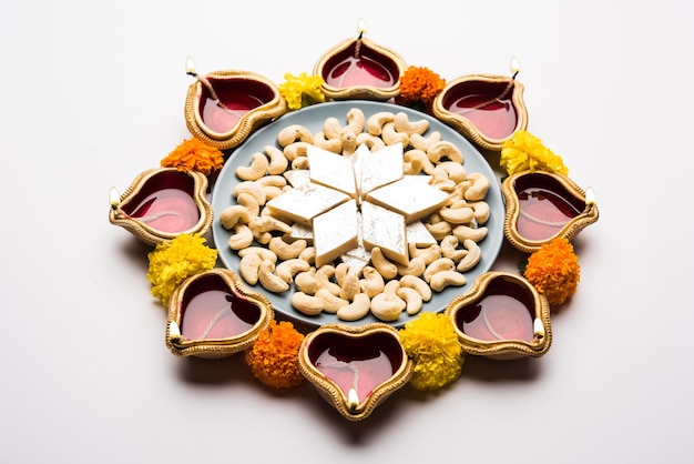 Diwali Food Rangoli mit Kaju Katli süß zusammen mit Clay Diya oder Lampe und Ringelblumen in kreisförmigem Muster angeordnet, selektiver Fokus