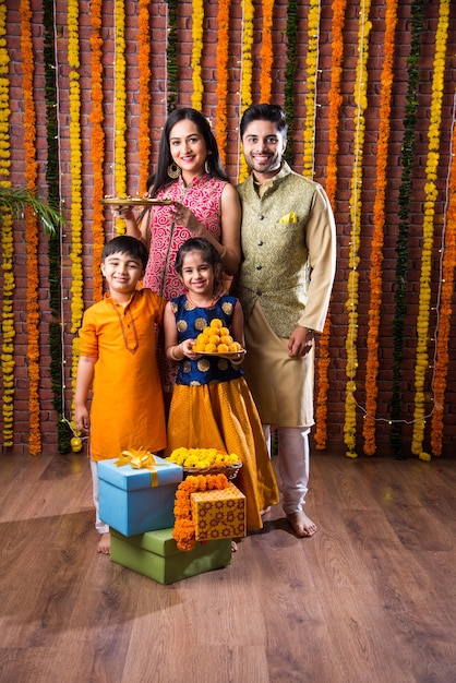 Diwali- oder Rakshabandhan-Feier - Indische junge vierköpfige Familie, die das Deepavali- oder Bhai-Dooj-Festival mit süßem Laddoo, Öllampe oder Diya und Geschenkboxen feiert, Essen isst oder Selfies macht