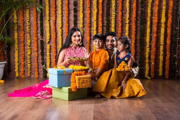 Diwali- oder Rakshabandhan-Feier - Indische junge vierköpfige Familie, die das Deepavali- oder Bhai-Dooj-Festival mit süßem Laddoo, Öllampe oder Diya und Geschenkboxen feiert, Essen isst oder Selfies macht