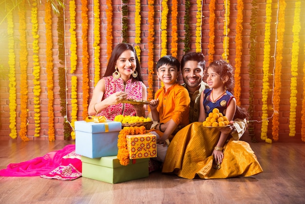 Diwali- oder Rakshabandhan-Feier - Indische junge vierköpfige Familie, die das Deepavali- oder Bhai-Dooj-Festival mit süßem Laddoo, Öllampe oder Diya und Geschenkboxen feiert, Essen isst oder Selfies macht