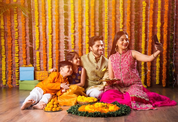 Diwali- oder Rakshabandhan-Feier - Indische junge vierköpfige Familie, die das Deepavali- oder Bhai-Dooj-Festival mit süßem Laddoo, Öllampe oder Diya und Geschenkboxen feiert, Essen isst oder Selfies macht