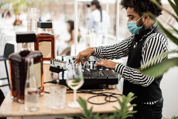 Dj africano tocando música em um bar de coquetéis ao ar livre enquanto usa máscara de segurança - foco principal na parte superior