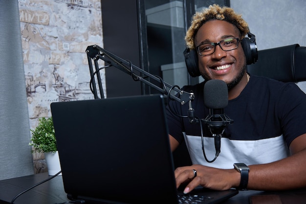 DJ de rádio com penteado afro e óculos apresenta um programa popular para ouvintes O podcaster preto alegre olha para a câmera sentado no estúdio com um microfone profissional e fones de ouvido