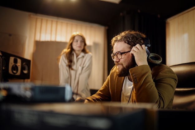 DJ masculino e cantora, gravando o interior do estúdio no fundo. Sintetizador e mixer de áudio, local de trabalho do músico, processo criativo