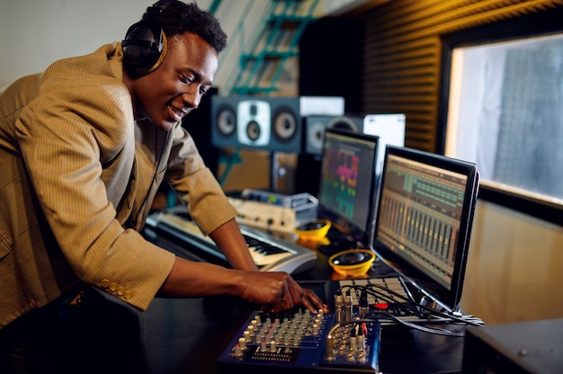 DJ masculino em fones de ouvido, ouvindo um disco no console de mixagem, gravando o interior do estúdio no fundo. Sintetizador e mixer de áudio, local de trabalho do músico, processo criativo