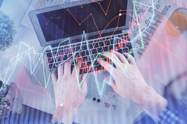 Doble exposición de las manos del hombre escribiendo en el teclado de la computadora portátil y dibujo de hologramas de gráficos de divisas Vista superior Concepto de mercados financieros