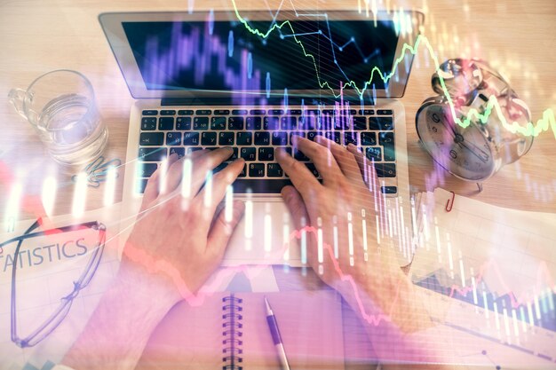 Doble exposición de las manos del hombre escribiendo en el teclado de la computadora portátil y dibujo de hologramas de gráficos de divisas Vista superior Concepto de mercados financieros