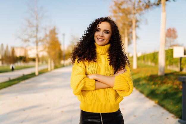 Dobrando no peito as mãos de uma garota em um suéter amarelo brilhante, bonito sorrindo fica no parque