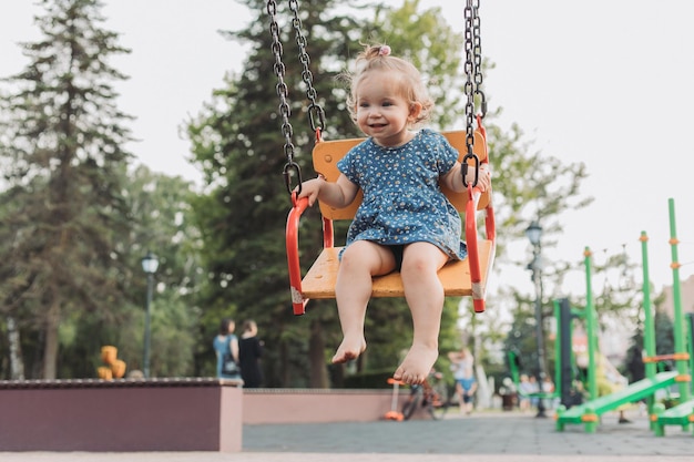 Doce bebê em um vestido azul balança em um balanço em um playground no estilo de vida do parque