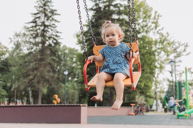 Doce bebê em um vestido azul balança em um balanço em um playground no estilo de vida do parque