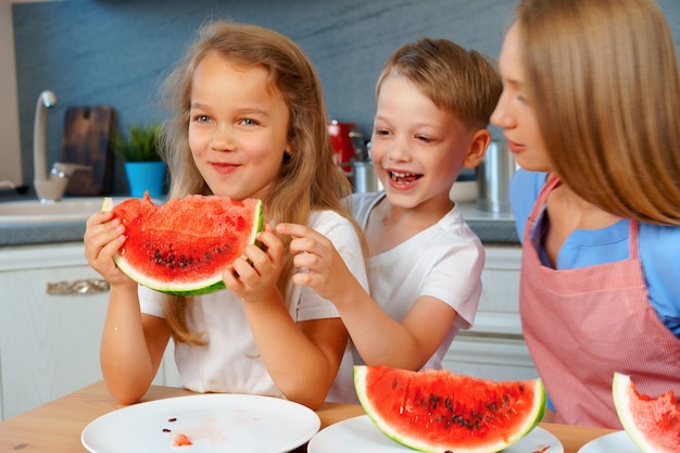 Doce família, mãe e filhos comendo melancia na cozinha se divertindo