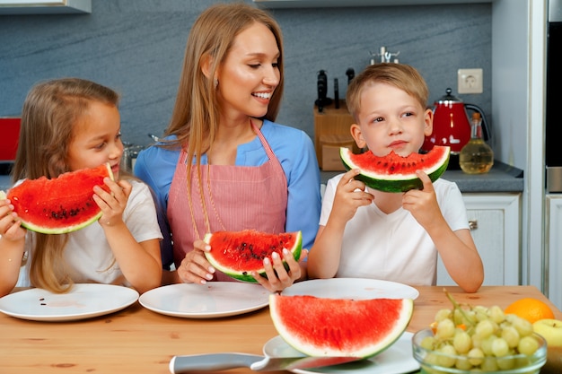 Doce família, mãe e filhos comendo melancia na cozinha se divertindo