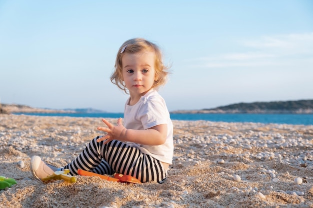 Doce garotinha caucasiana com cabelo loiro encaracolado brincar na praia de areia com pedras pelo mar azulFelizbonitoadorávelbebê infantilcriança passar o tempo de fériasférias ao ar livre à beira-mar