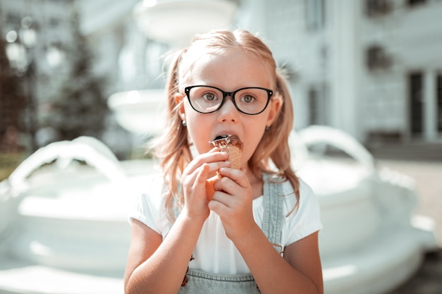 Doce infância. menina séria de óculos comendo sua casquinha de sorvete de chocolate do lado de fora, segurando-a com as duas mãos.