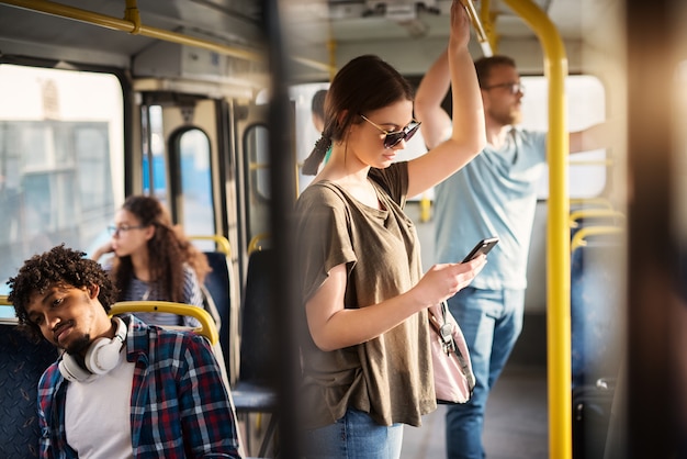 Doce menina com óculos de sol em usar o telefone em pé em um ônibus.