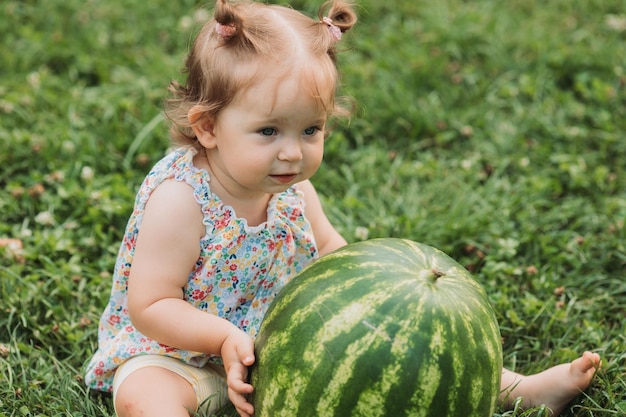 Doce menina com uma enorme melancia está sentado em um gramado verde criança brinca ao ar livre estilo de vida