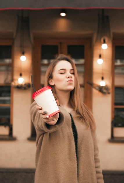 Doce menina mostra um beijo e uma xícara de café nas mãos, veste um casaco bege, fica contra o fundo da parede do restaurante