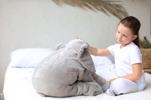 Doce menina sentada em sua cama em casa com o elefante de brinquedo.
