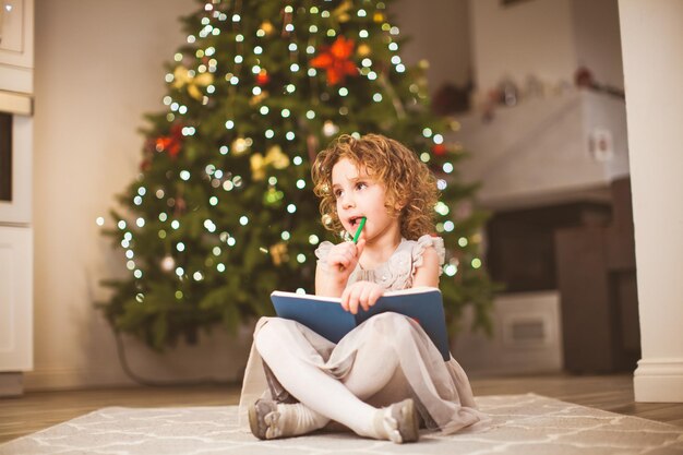 Doce menina sonhadora com cabelos cacheados, vestido cinza, sentado na frente da árvore de Natal decorada com as pernas cruzadas Garota pensando em carta para o Papai Noel segurando caderno e lápis