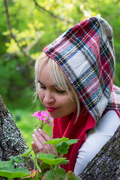 Doce mulher com capuz na floresta, cheirando o rico aroma de uma linda flor rosa.