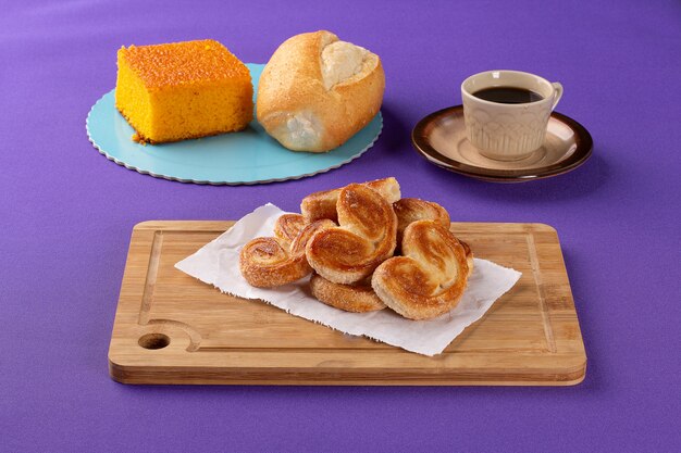 Doces de padaria em uma placa de madeira com um bolo de cenoura e pão francês ao lado de uma xícara