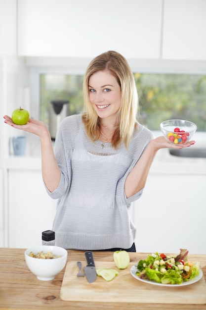 Doces e mulheres escolhidos decidem sobre comida saudável em uma cozinha para uma dieta e segurando uma maçã e doces em casa O retrato sorri uma pessoa feliz do sexo feminino com frutas para nutrição como refeição de almoço