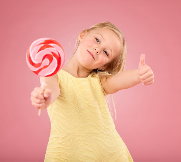 Doces polegares para cima e pirulito com retrato de menina em estúdio para festa de açúcar e comida de carnaval isolada em fundo rosa Bonito positivo e jovem com criança ou comendo lanche para brincar e tratar