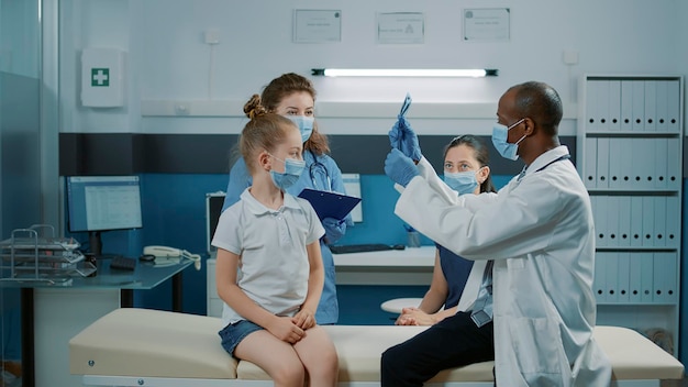 Doctor analizando los resultados de la radiografía con la niña y el padre en la visita de control. Médico explicando el diagnóstico de exploración de rayos X a un paciente pequeño y a la madre en la cita durante la pandemia de covid 19.