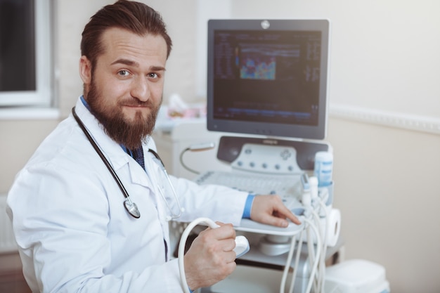 Doctor barbudo amigable sonriendo a la cámara, sosteniendo un escáner de ultrasonido en la mano