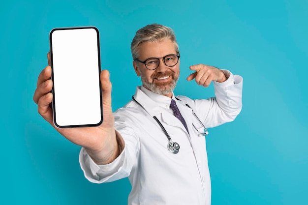 Doctor caucásico canoso sonriente que muestra el teléfono con la pantalla en blanco