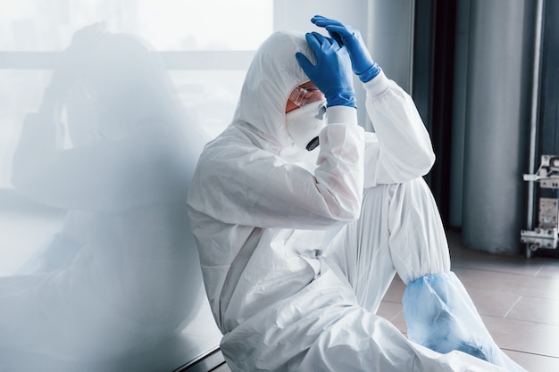 Doctor científico cansado en bata de laboratorio, gafas y máscara defensiva se sienta y toma un descanso