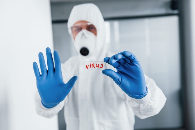 Doctor científico masculino en bata de laboratorio, gafas defensivas y máscara sostiene vidrio con palabra de virus