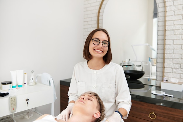 Doctor-cosmetólogo sonriente sentado cerca de una mujer cliente en la clínica de spa de belleza contemporánea.