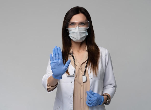 Doctor / enfermera sonriendo detrás de la máscara del cirujano. Modelo de mujer caucásica joven en matorral médico blanco y guantes desechables mostrando gesto de parada