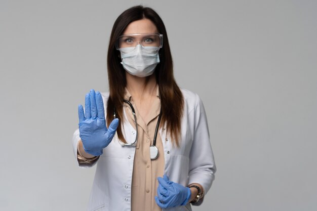 Doctor / enfermera sonriendo detrás de la máscara del cirujano. Modelo de mujer caucásica joven en matorral médico blanco y guantes desechables mostrando gesto de parada