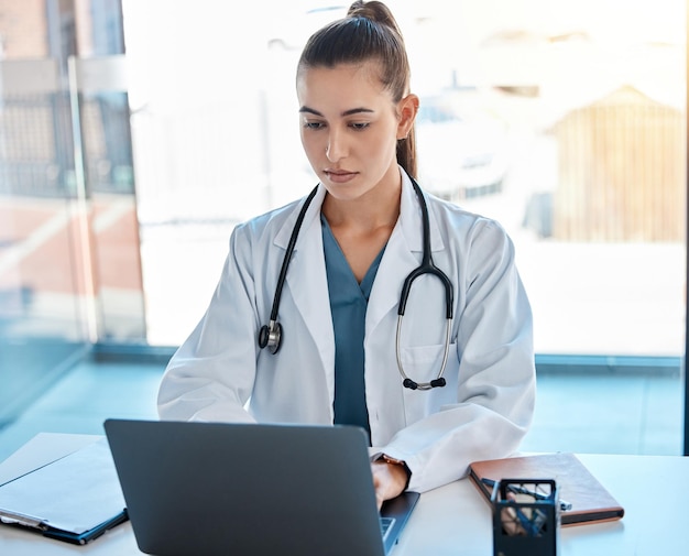 Doctor escribiendo en una computadora portátil en una oficina escribiendo una propuesta o en una videollamada usando telemedicina joven profesional de la salud o médico de cabecera usando internet para investigación o consulta en línea