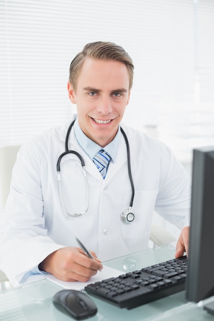 Doctor escribiendo una nota mientras usa la computadora en el consultorio médico