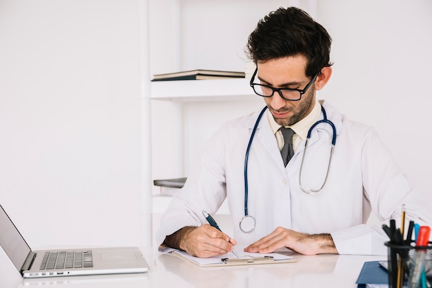 Doctor escribiendo en el portapapeles con el portátil en el escritorio