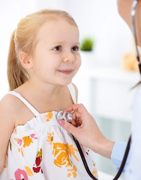 Doctor examinando a un niño con estetoscopio en una clínica soleada. Una feliz paciente sonriente vestida con un vestido de color brillante está en la inspección médica habitual.