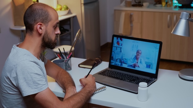 Doctor explicando el diagnóstico durante una videoconferencia y paciente escribiendo en un cuaderno. Médico que consulta al paciente enfermo desde la oficina del hospital durante el examen virtual trabajando horas extraordinarias.