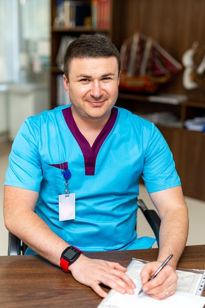 Doctor feliz escribiendo algo y mirando a la cámara mientras está sentado en la mesa en su gabinete Hombre guapo en uniforme médico Concepto de medicina