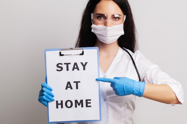 Doctor en guantes, máscara y gafas sostiene la hoja con texto quedarse en casa.