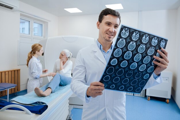 Doctor guapo alegre agradable mirando la imagen de rayos X y estar de buen humor mientras está de pie en el laboratorio médico