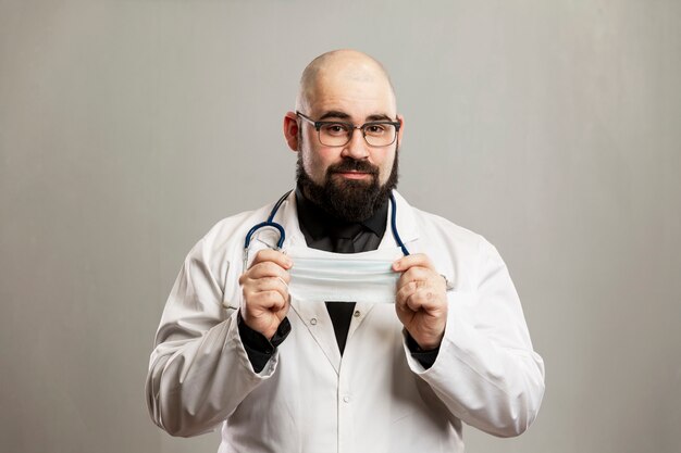 Doctor hombre en una bata blanca con una máscara médica en sus manos. Pared gris.